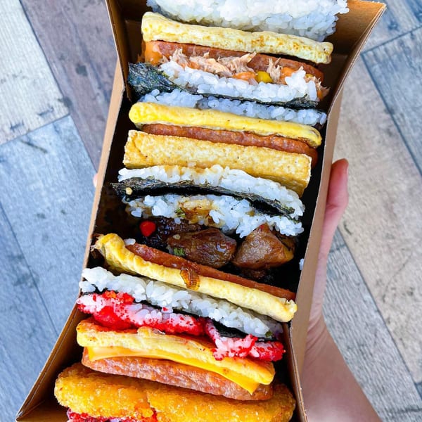 A hand holds a long box filled with various Supamu Okinawa Onigiri, featuring rice, spam, egg, and other fillings.