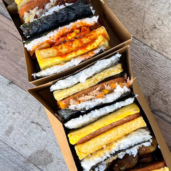 Two open cardboard boxes filled with various Supamu Okinawa Onigiri on a wooden surface.