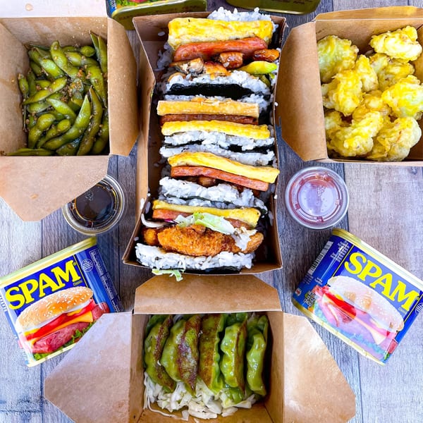 An overhead view of Supamu Okinawa Onigiri's takeout boxes filled with spam musubi, edamame, tempura, and gyoza.